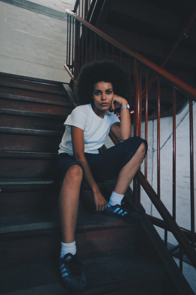 Mila Smith sits on a wooden staircase, leaning on one arm, wearing a white t-shirt, dark shorts, and sneakers in a softly lit interior.