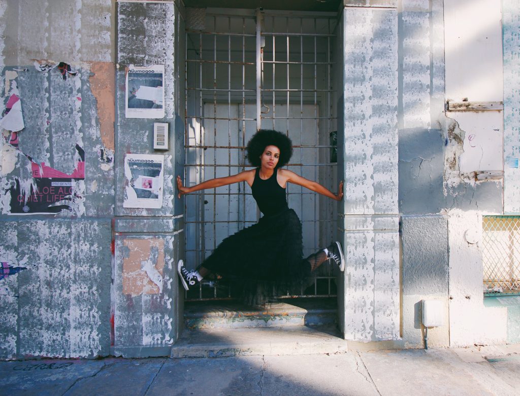 Mila Smith poses in a wide stance in a weathered doorway, wearing a black dress and sneakers, framed by textured urban walls.