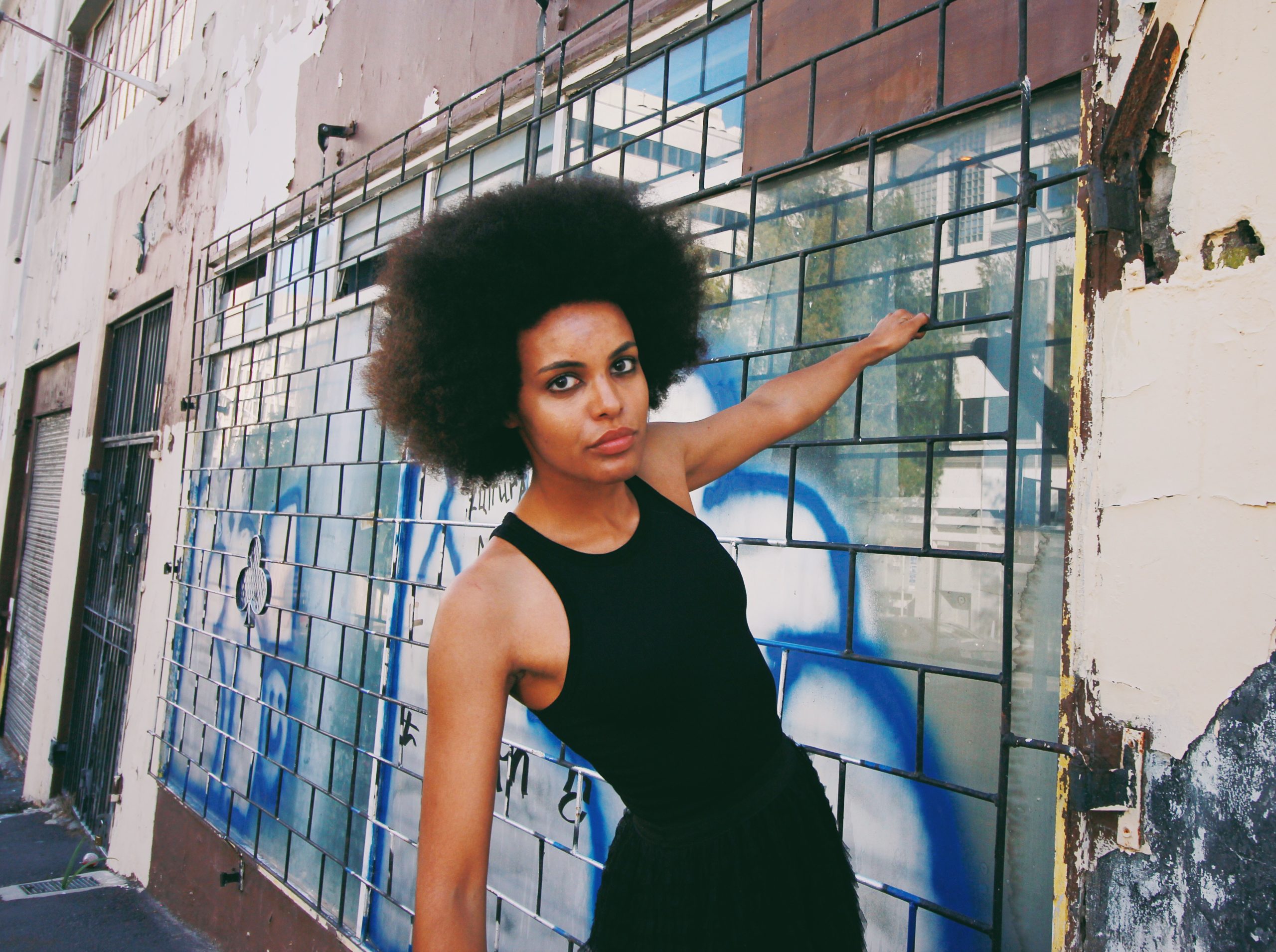 Mila Smith leans against a tiled exterior wall with graffiti, wearing a black sleeveless top and looking directly at the camera.