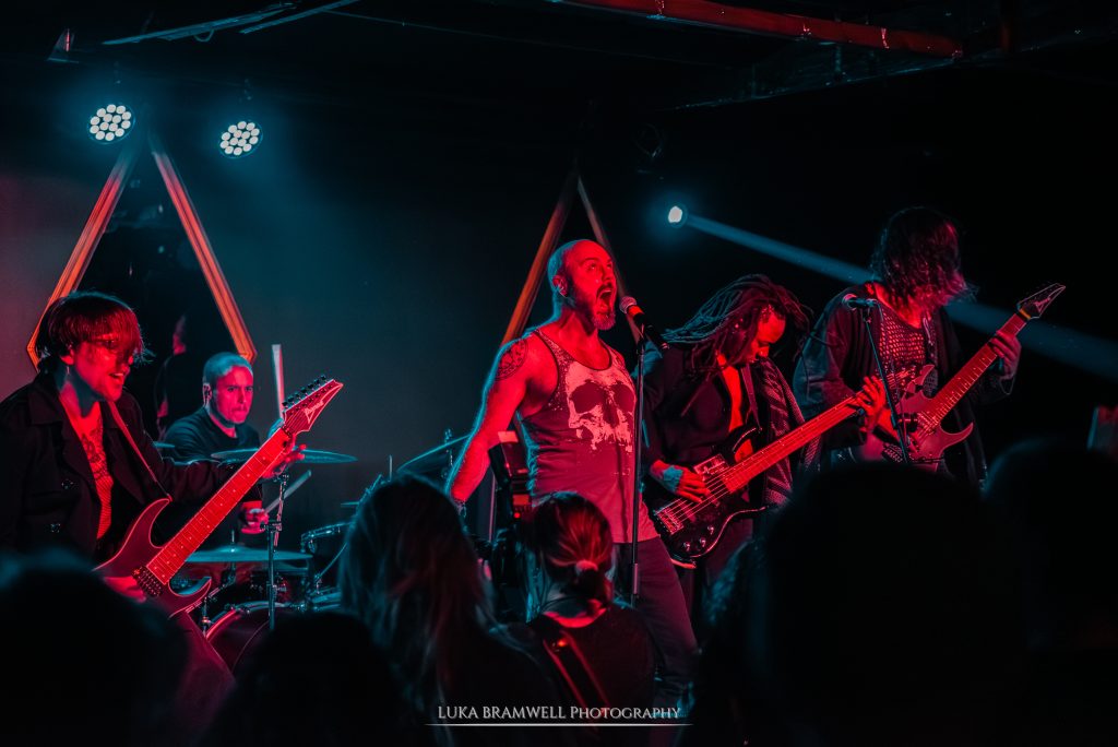 A wide live performance shot of the full Dreadlines lineup on stage. The vocalist stands center-stage in a skull tank top, shouting into the microphone, flanked by two guitarists, a bassist with long dreadlocks, and a drummer in the background. The scene is bathed in contrasting teal and red stage lighting, creating a high-energy atmosphere.