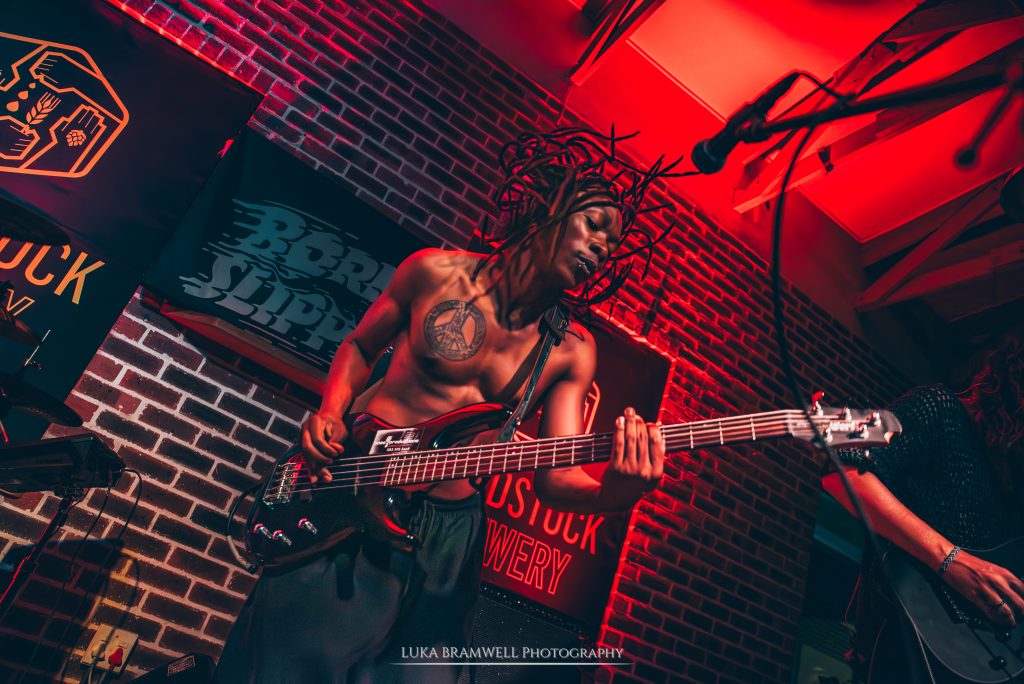 A high-energy live performance shot of a member of the band Dreadlines playing a black electric bass guitar on stage. The musician, featuring long dreadlocks and a peace sign chest tattoo, is captured mid-performance under intense red stage lighting. In the background, a brick wall is adorned with a "Woodstock Brewery" banner.