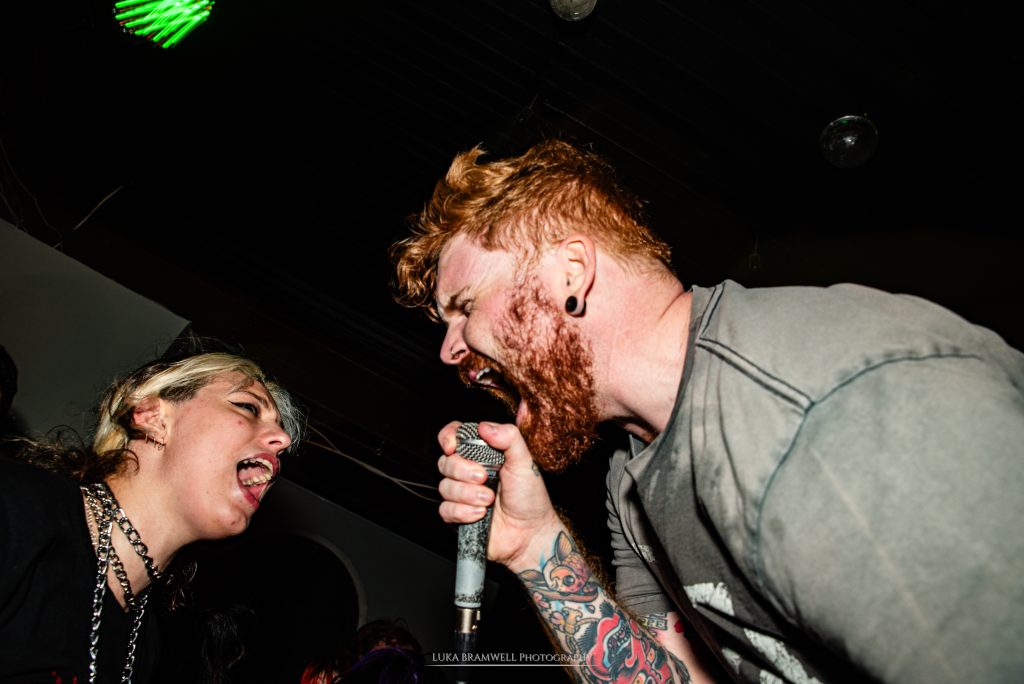 A dynamic close-up of The Talon's vocalist, Scott Miller, screaming into a microphone during a concert. He is in profile, showing his red beard and tattooed arm, while a fan on the left screams along with him, inches from the mic. The shot is taken from a low angle against a dark background, emphasizing the raw, visceral connection between the performer and the audience.