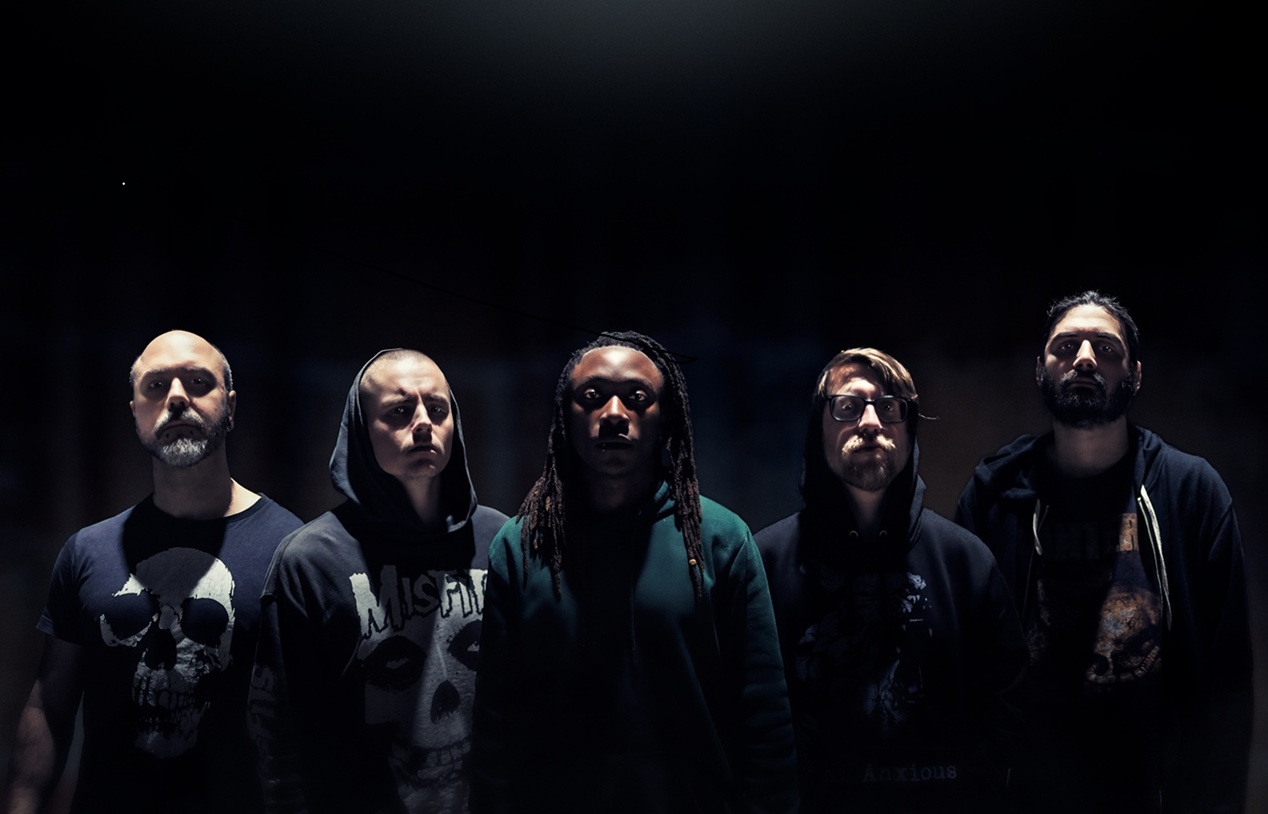 A moody, high-contrast promotional portrait of the five members of Dreadlines. The band is arranged in a horizontal line against a pitch-black background, illuminated by a single harsh overhead spotlight that creates dramatic shadows. The members are dressed in dark hoodies and t-shirts—including Misfits and skull graphics—staring intensely at the camera. The central figure features long dreadlocks, grounding the composition.