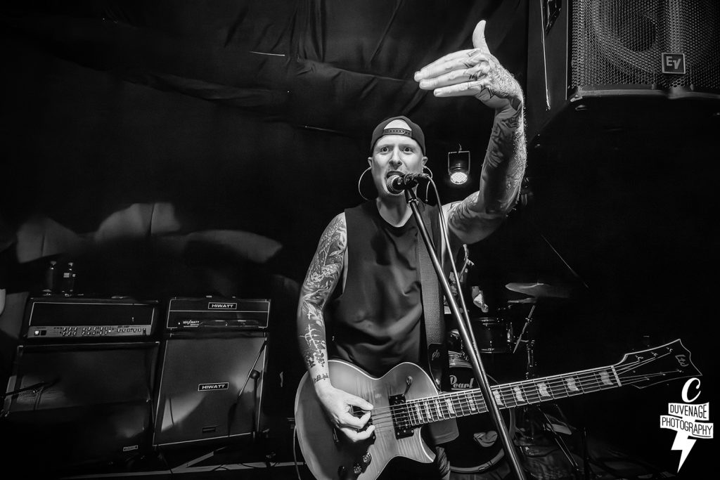 A high-energy black and white live performance photo of GRIFT. The guitarist, sporting tattoos and a backwards cap, sings into a microphone while playing an LTD electric guitar. Two Hiwatt amplifiers are visible in the background of the dark stage.
