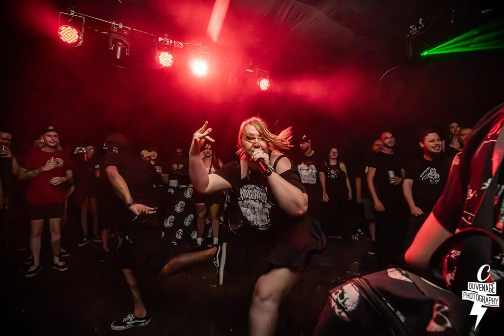 GRIFT performing live at a crowded venue under intense red stage lighting. The vocalist is captured in motion, singing passionately with one hand raised in a gesture, while a fan in the background appears to be dancing or moshing.