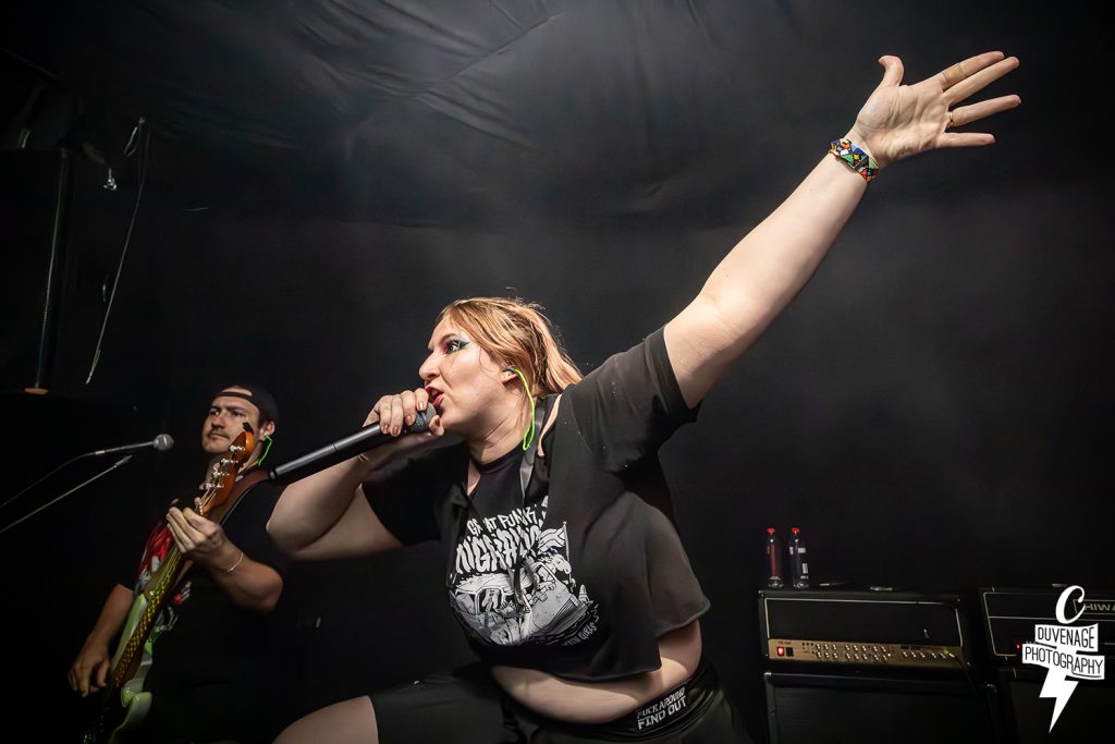 An action-oriented live shot of the band GRIFT. The vocalist, wearing colorful eye makeup and a graphic t-shirt, leans forward with one arm outstretched toward the ceiling. A bassist is visible in the background beside a stack of Hiwatt amplifiers.