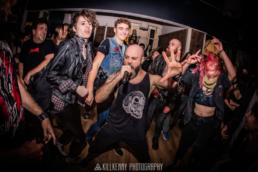 A dynamic wide-angle "fisheye" photograph showing the lead vocalist of Dreadlines performing in the middle of a crowded, energetic mosh pit. The vocalist, wearing a black tank top with a skull graphic, screams into a microphone while surrounded by fans in a dimly lit venue. The image captures the raw intensity of a live Dreadlines show.