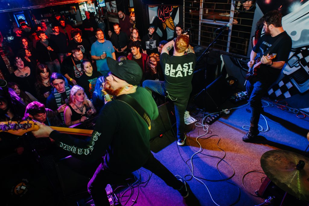 A high-angle, wide shot from behind The Talon looking out into the crowd during a live performance. The guitarist and bassist are in the foreground, with the lead singer wearing a "BLAST BEATS" t-shirt facing a packed, diverse audience. The venue is bathed in blue and red stage lights, capturing the intense energy and movement of the Cape Town hardcore scene.