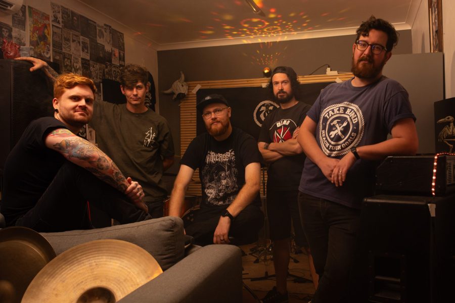 A wide-angle, promotional group shot of the five members of The Talon in a dimly lit rehearsal room. In the foreground, a member with red hair and arm tattoos sits on a couch near drum cymbals. Behind him, the other four members pose among musical equipment, including an amplifier and a drum kit. The background features walls covered in band posters and a disco ball casting warm, yellow light across the ceiling.