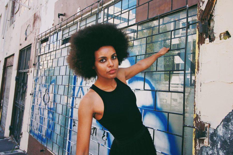 Mila Smith leans against a tiled exterior wall with graffiti, wearing a black sleeveless top and looking directly at the camera.