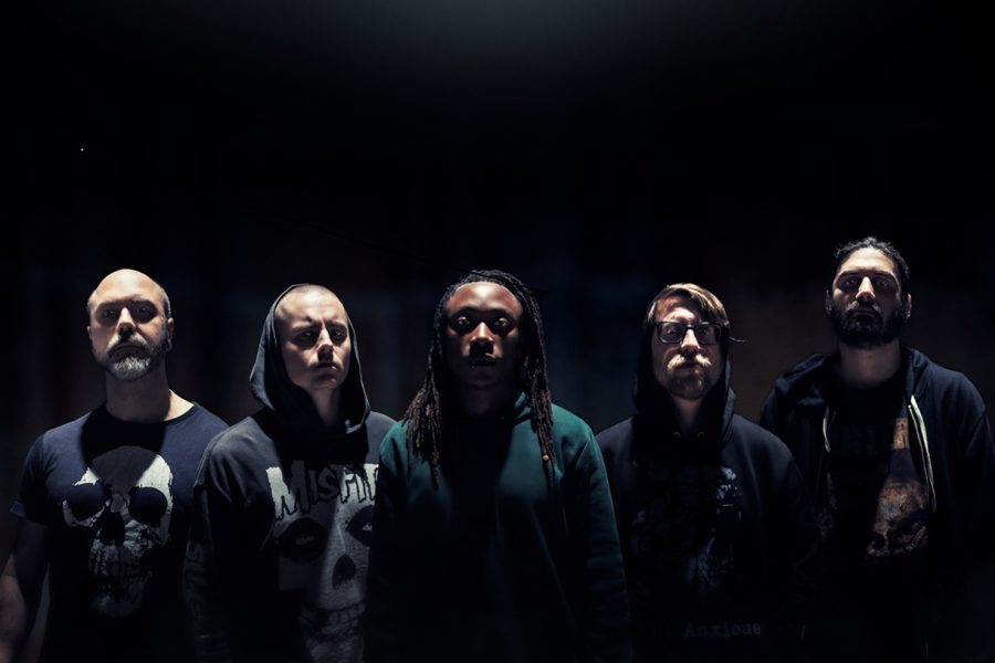 A moody, high-contrast promotional portrait of the five members of Dreadlines. The band is arranged in a horizontal line against a pitch-black background, illuminated by a single harsh overhead spotlight that creates dramatic shadows. The members are dressed in dark hoodies and t-shirts—including Misfits and skull graphics—staring intensely at the camera. The central figure features long dreadlocks, grounding the composition.